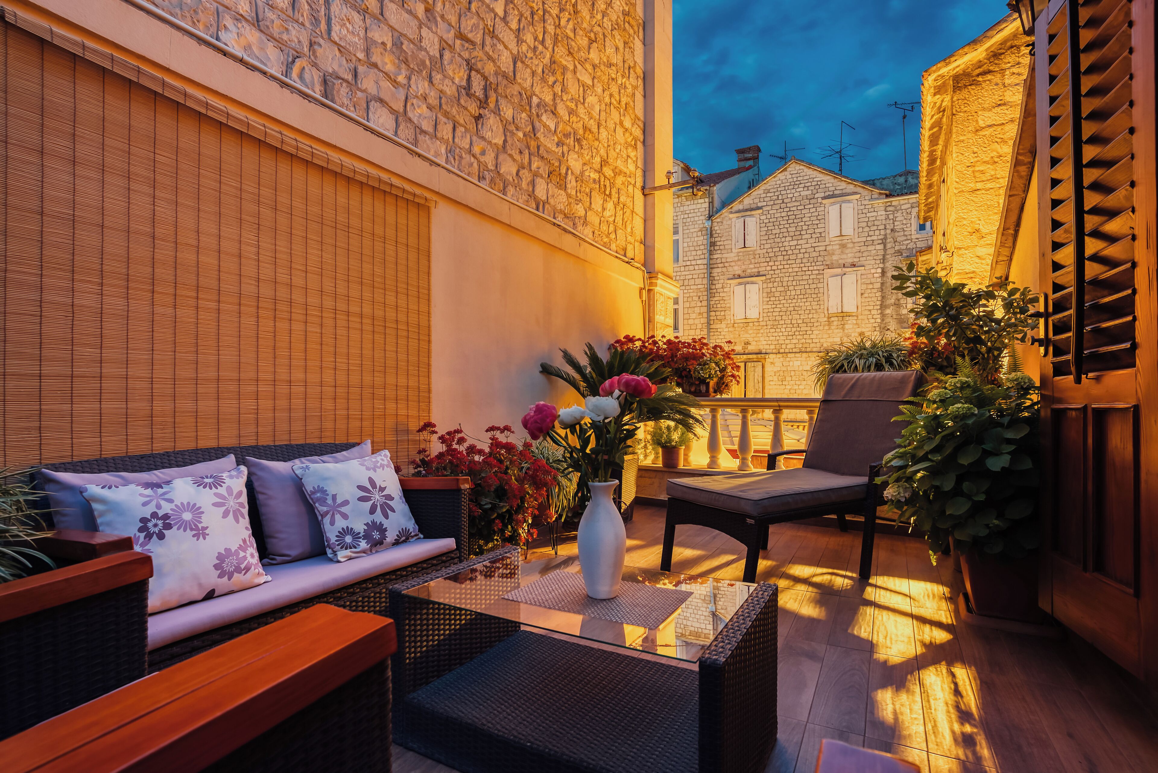 Une terrasse extérieure avec un canapé, des fauteuils et une table. On y trouve des plantes en pot et un vase rempli de fleurs.