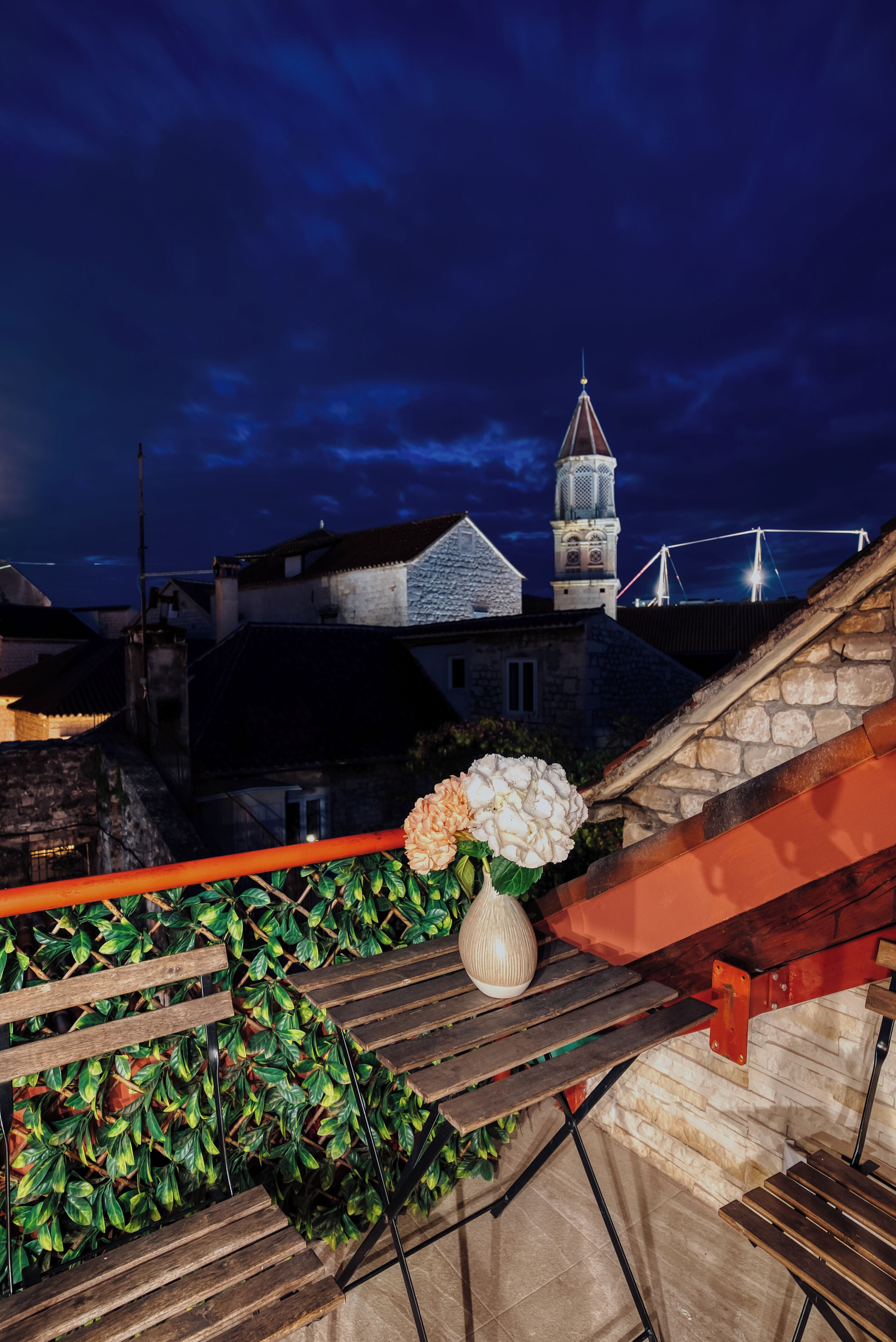 A rooftop terrace with a stone wall, wooden benches, a potted plant, and a view of a historic building with a tower.