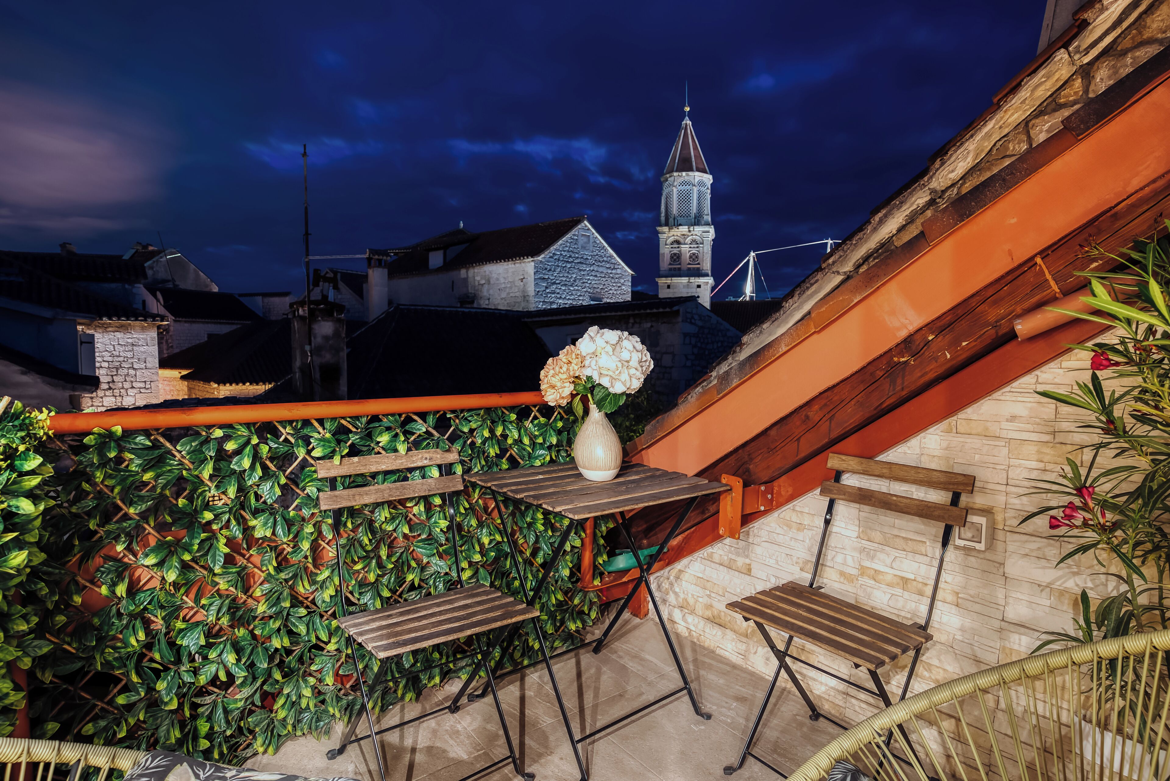 Un área de estar al aire libre con una mesa y sillas, un jarrón con flores y vistas a una torre de iglesia.