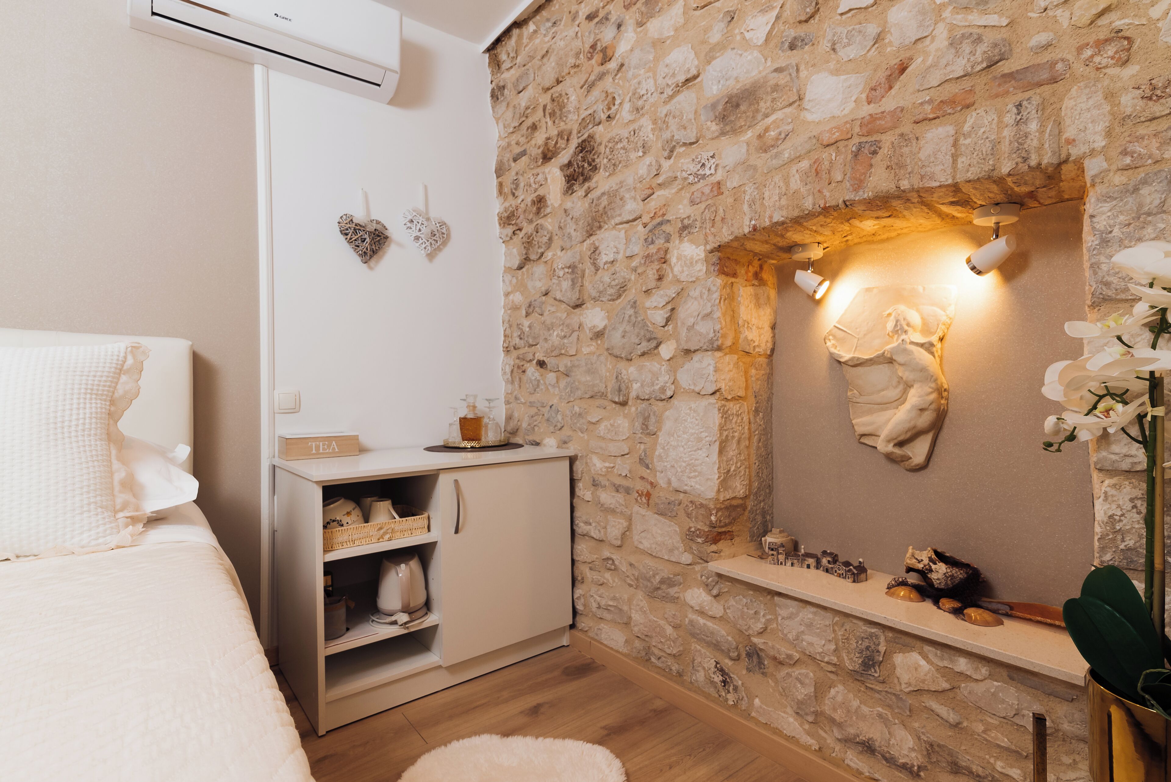 A bedroom with a stone wall, a bed with white bedding, a small cabinet, and a shelf with decorative items.