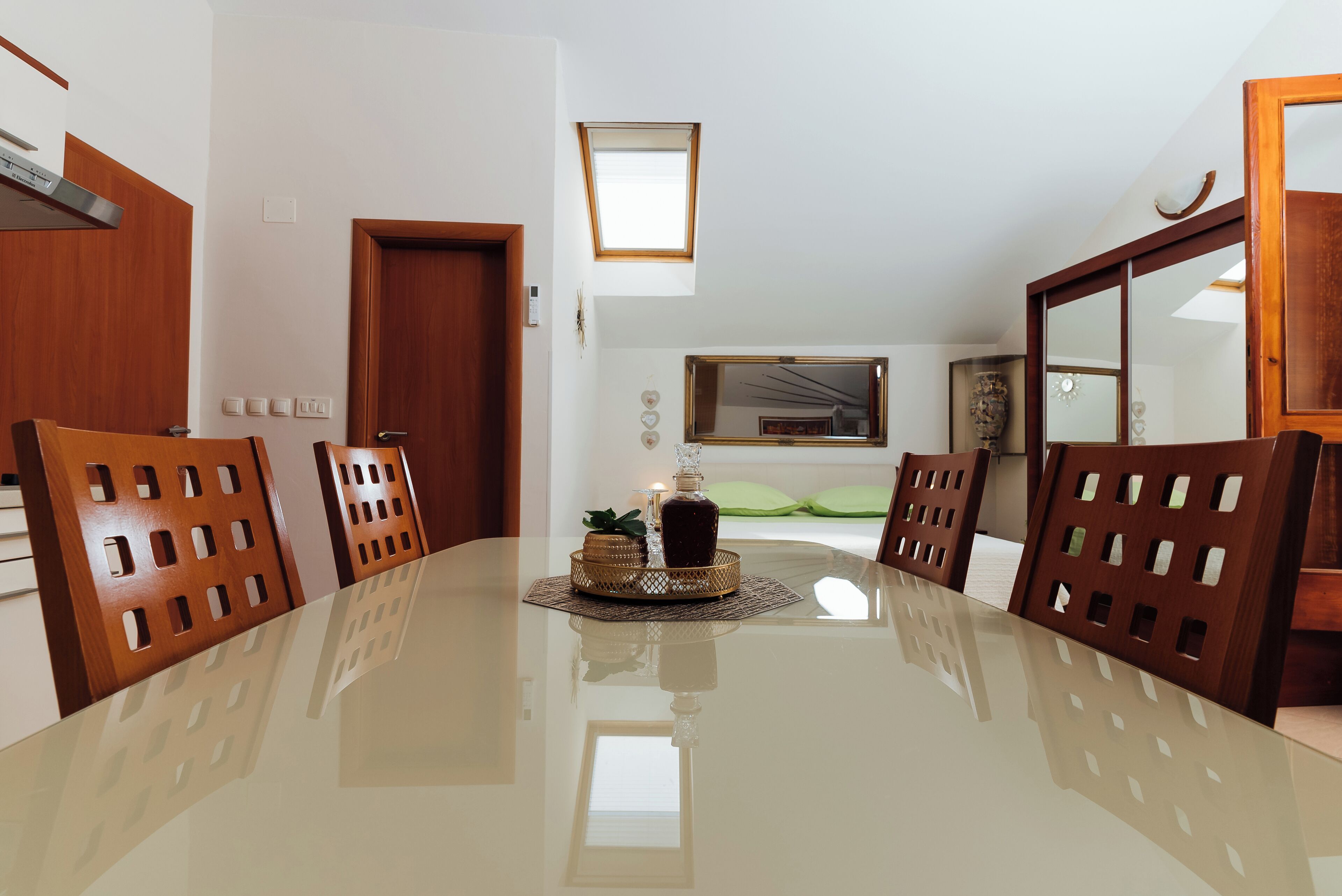 A dining area with a glass table, wooden chairs, a candle centerpiece, and a view of a room with a bed and a window.