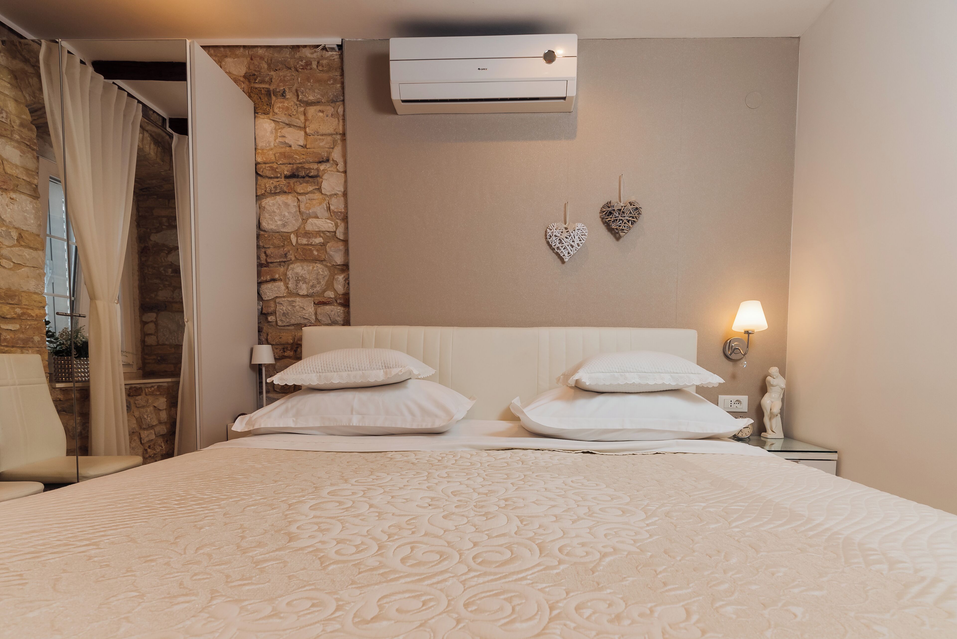 Ein Hotelzimmer mit Bett, weißer Bettwäsche, einer Steinwand, einer Klimaanlage, einem Nachttisch mit Lampe und dekorativen herzförmigen Wandgestaltungen.