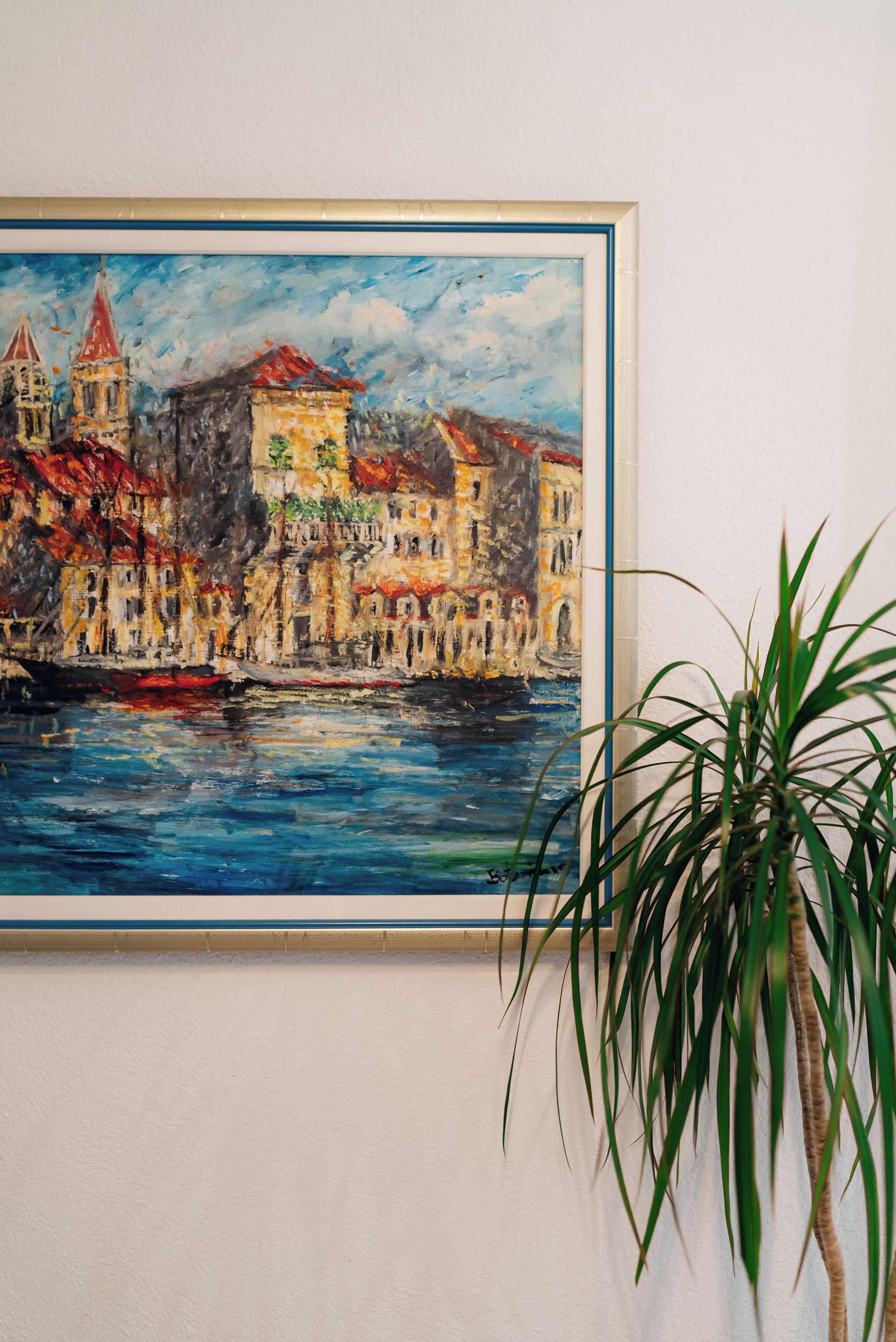 A framed painting of a colorful waterfront scene with buildings and a plant in the foreground.