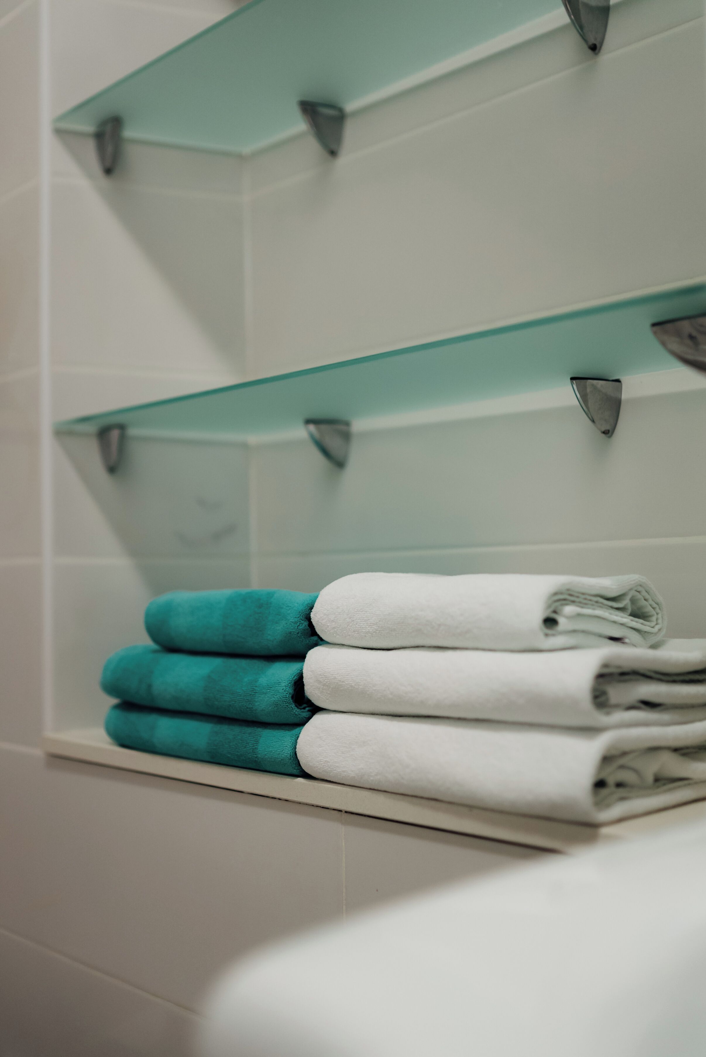 A bathroom with white tiled walls, a glass shelf with towels, and a white bathtub.