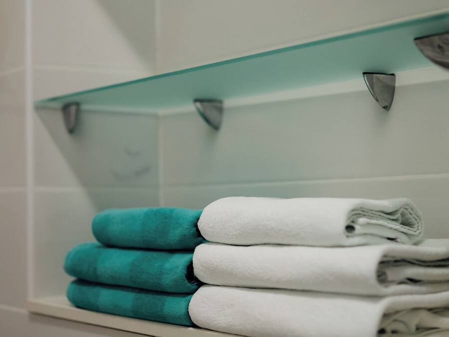 A bathroom with white tiled walls, a glass shelf with towels, and a white bathtub.
