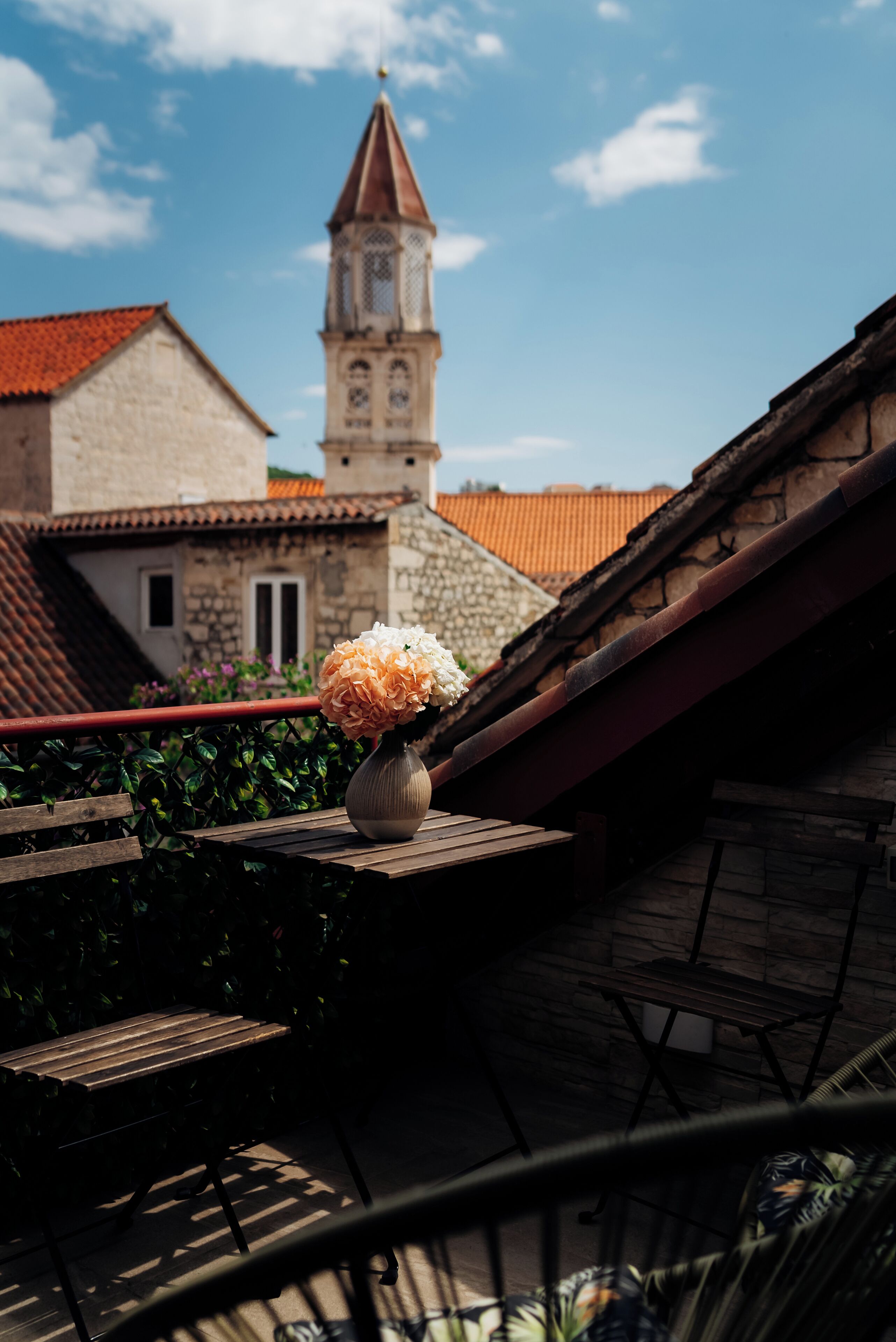 Un balcón con mesa de madera y un jarrón de flores, con vista a un edificio de piedra y tejado de tejas rojas y una torre.