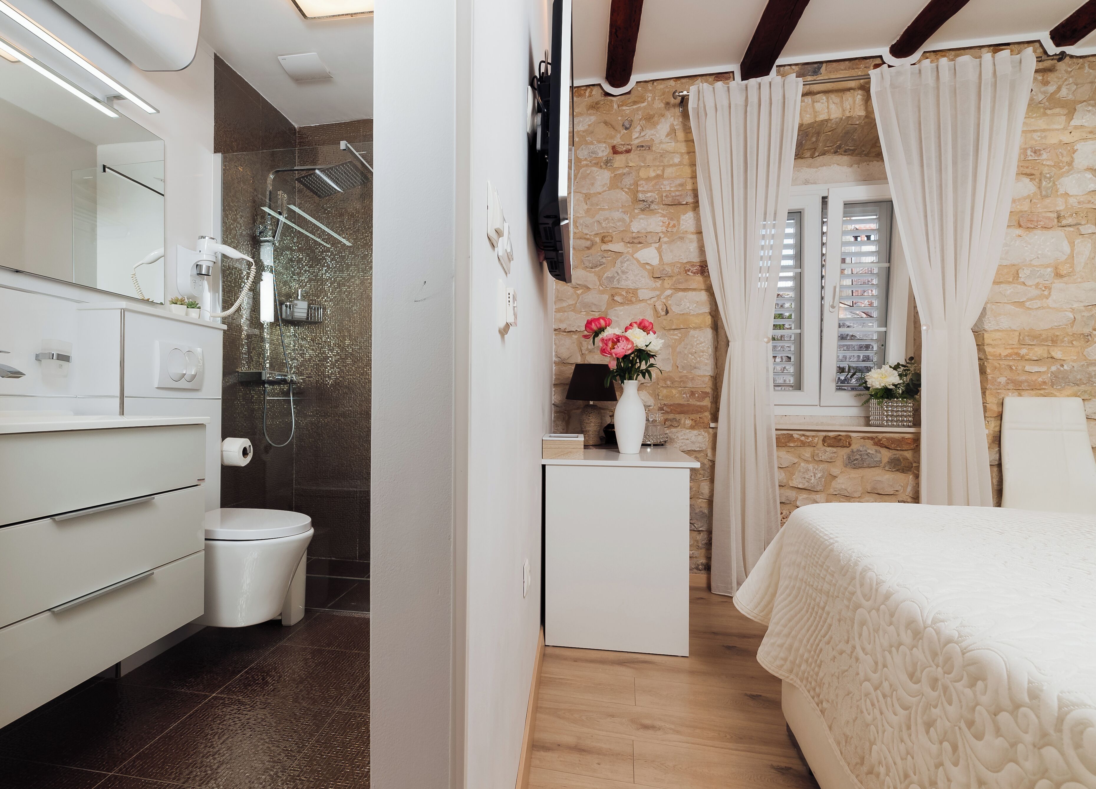 Een moderne badkamer met een douche, toilet en wastafel. Er is een bed met een decoratieve hoofdeinde en een raam met gordijnen.