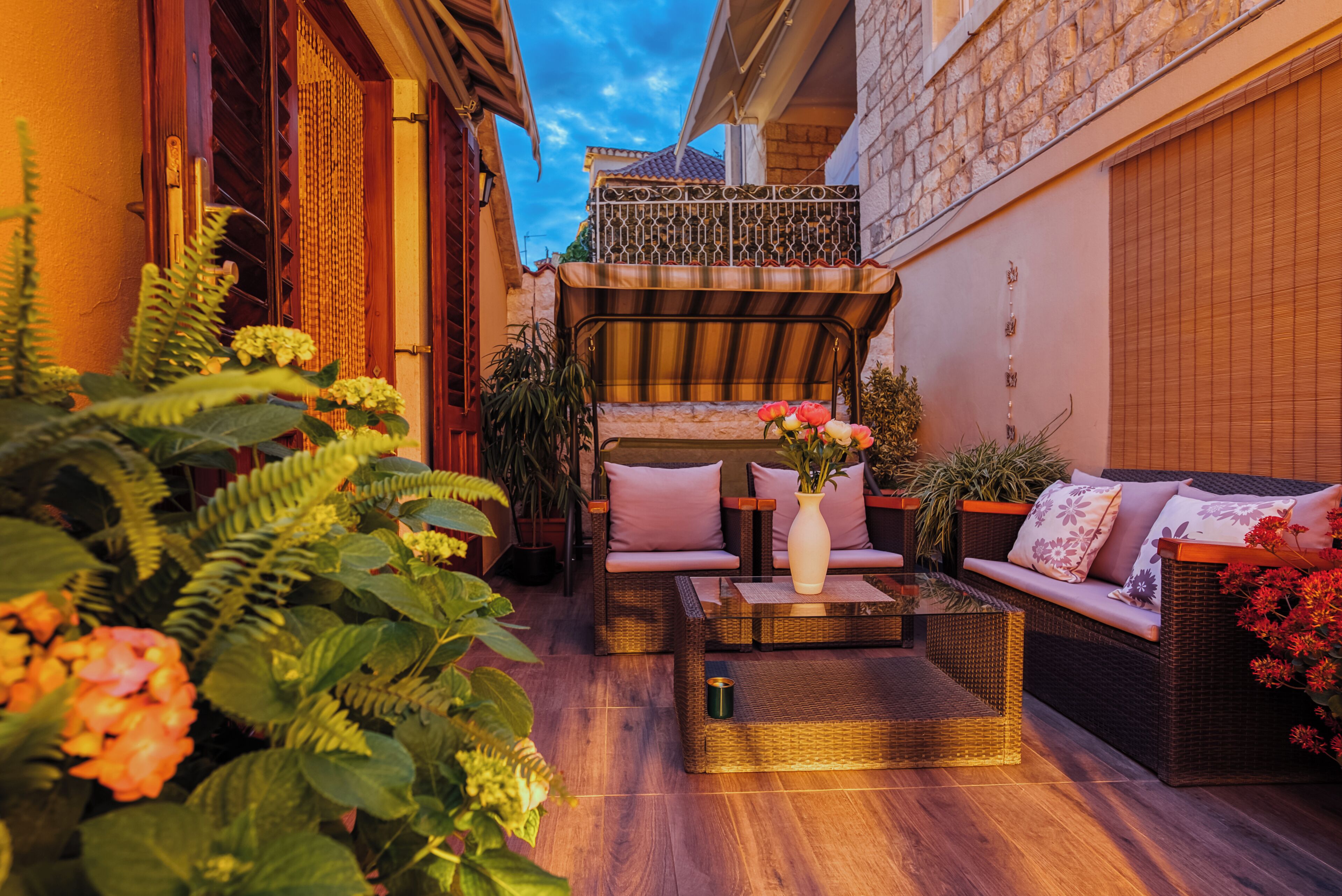 A cozy outdoor seating area with wicker furniture and a central coffee table, surrounded by lush plants and flowers.