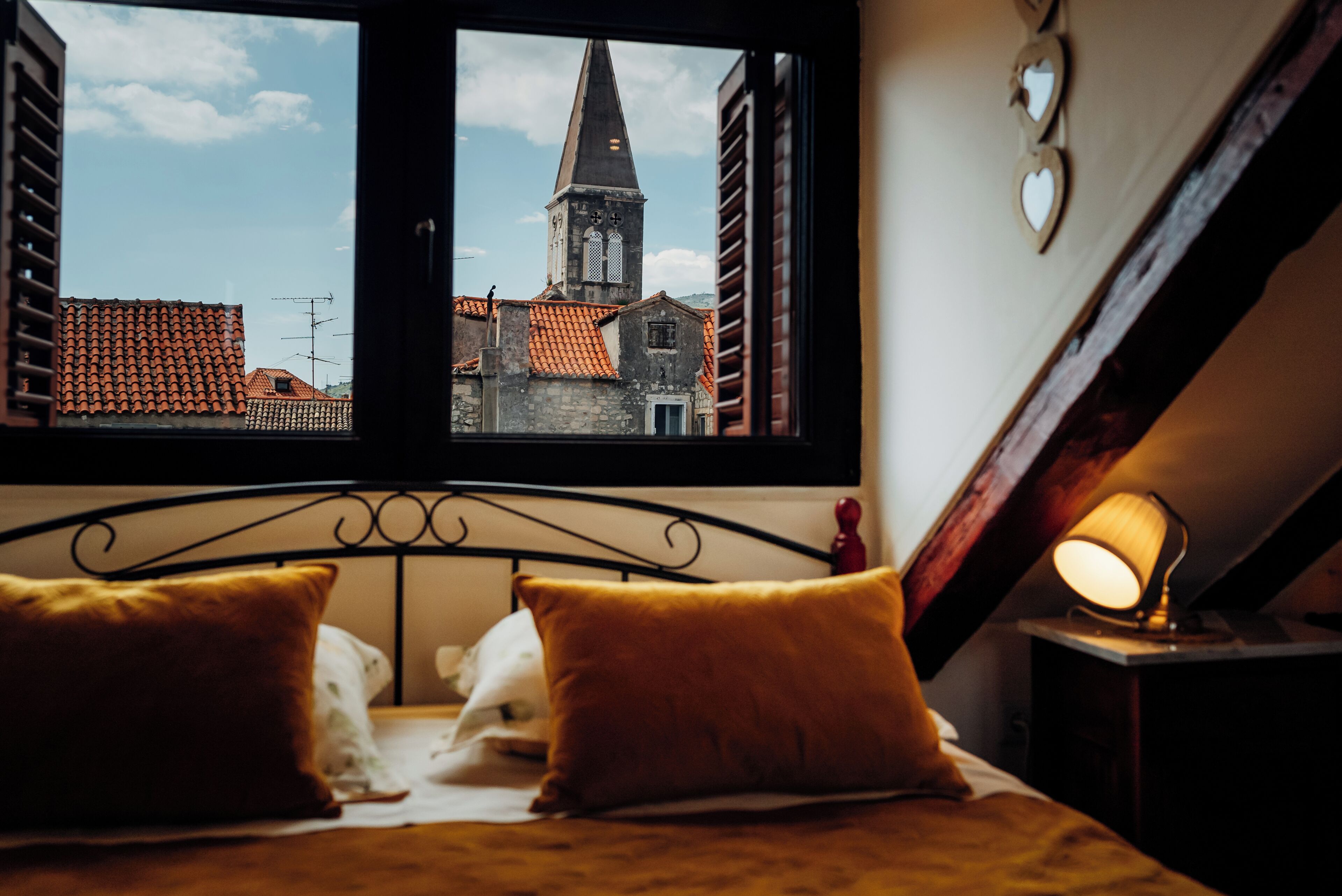 A bedroom with a view of a church steeple and red-tiled roofs through a window.