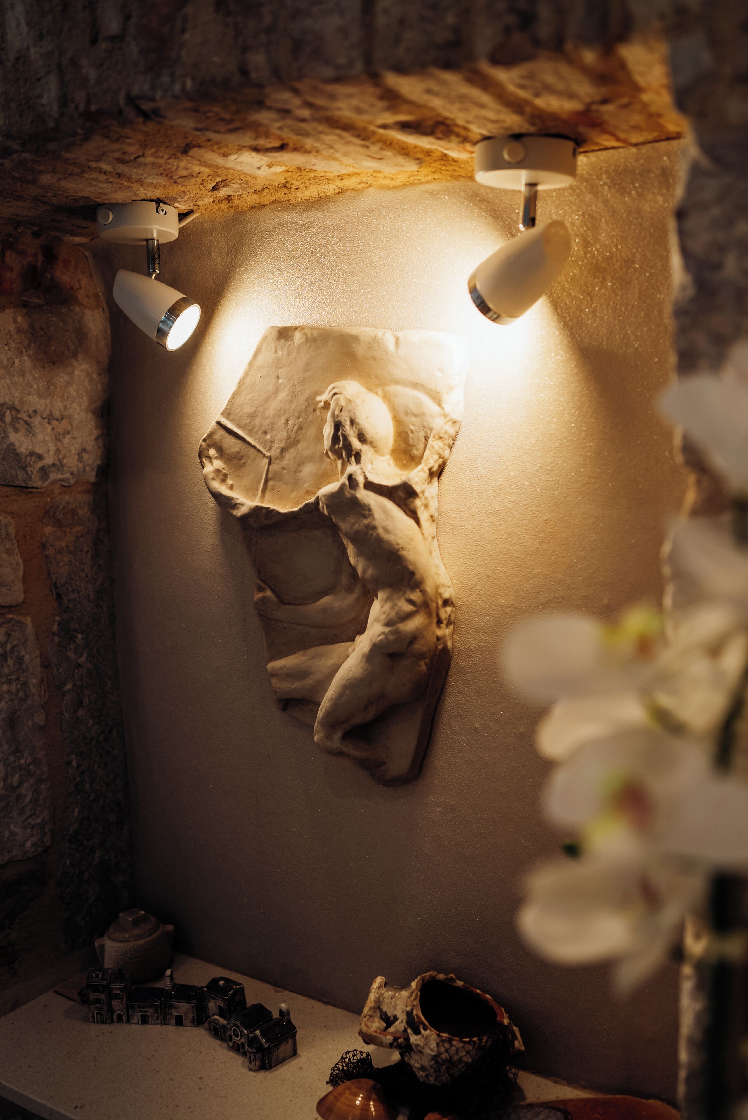 A dimly lit room with a stone wall, a small table with a skull and miniature buildings, and a light fixture above a wall-mounted object.