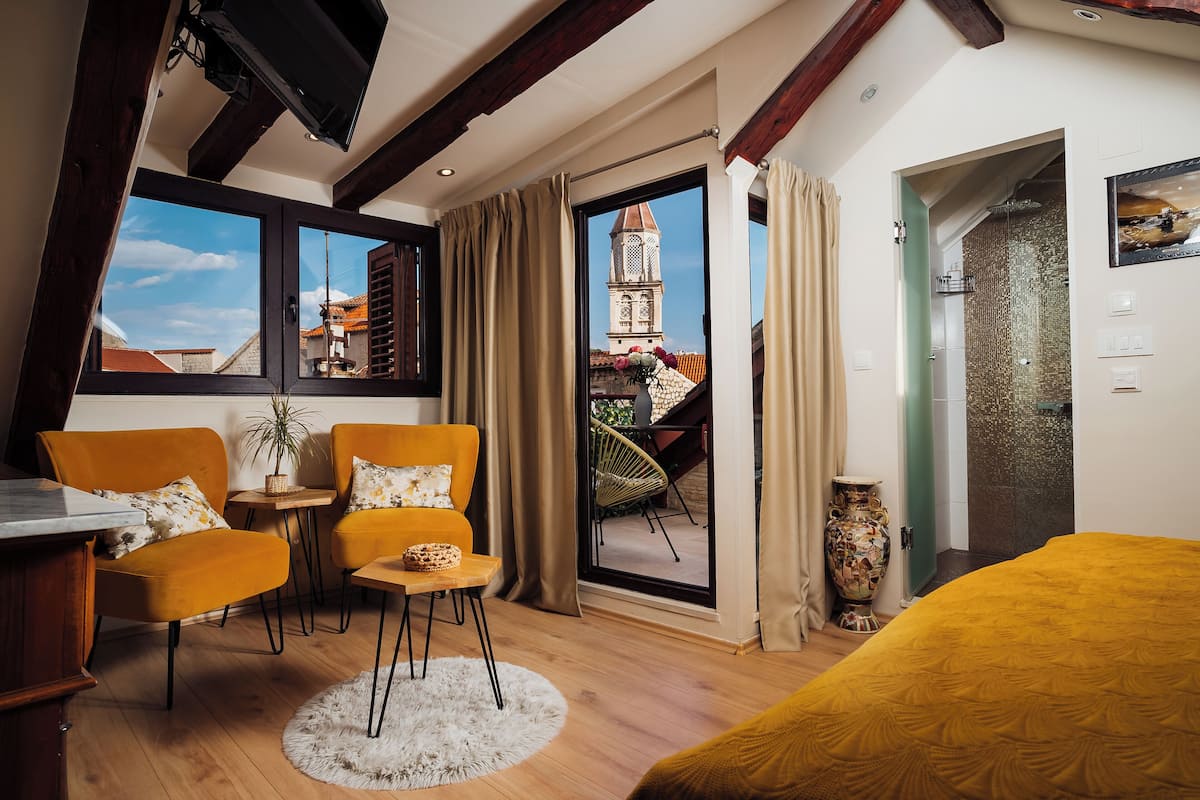 Ein Zimmer mit einem Bett, zwei Stühlen, einem kleinen Tisch, einer Zimmerpflanze und Blick auf ein Gebäude durch ein Fenster.