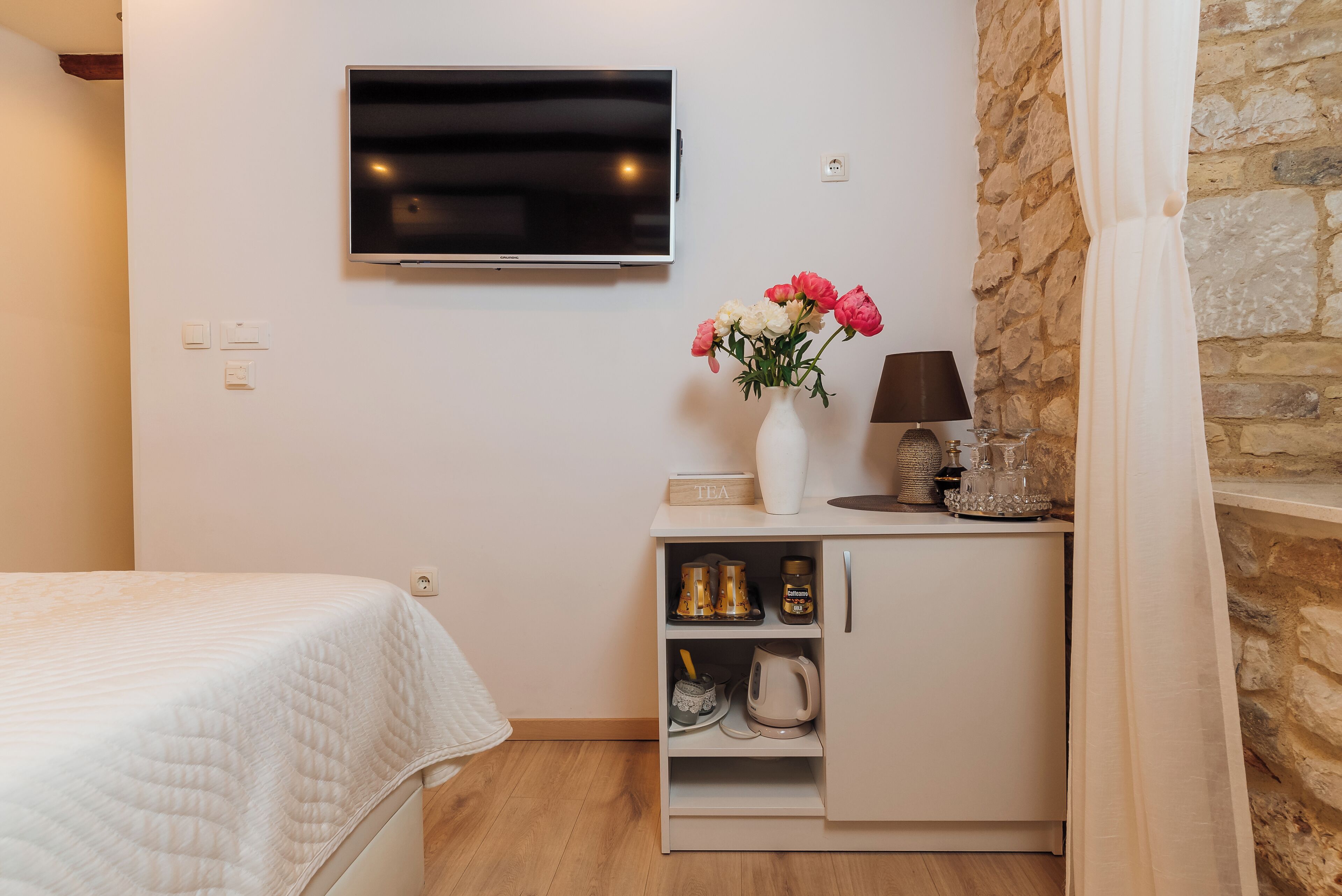 Une chambre avec un lit, un téléviseur fixé au mur, un petit meuble contenant du thé et du café, et un mur en pierre.