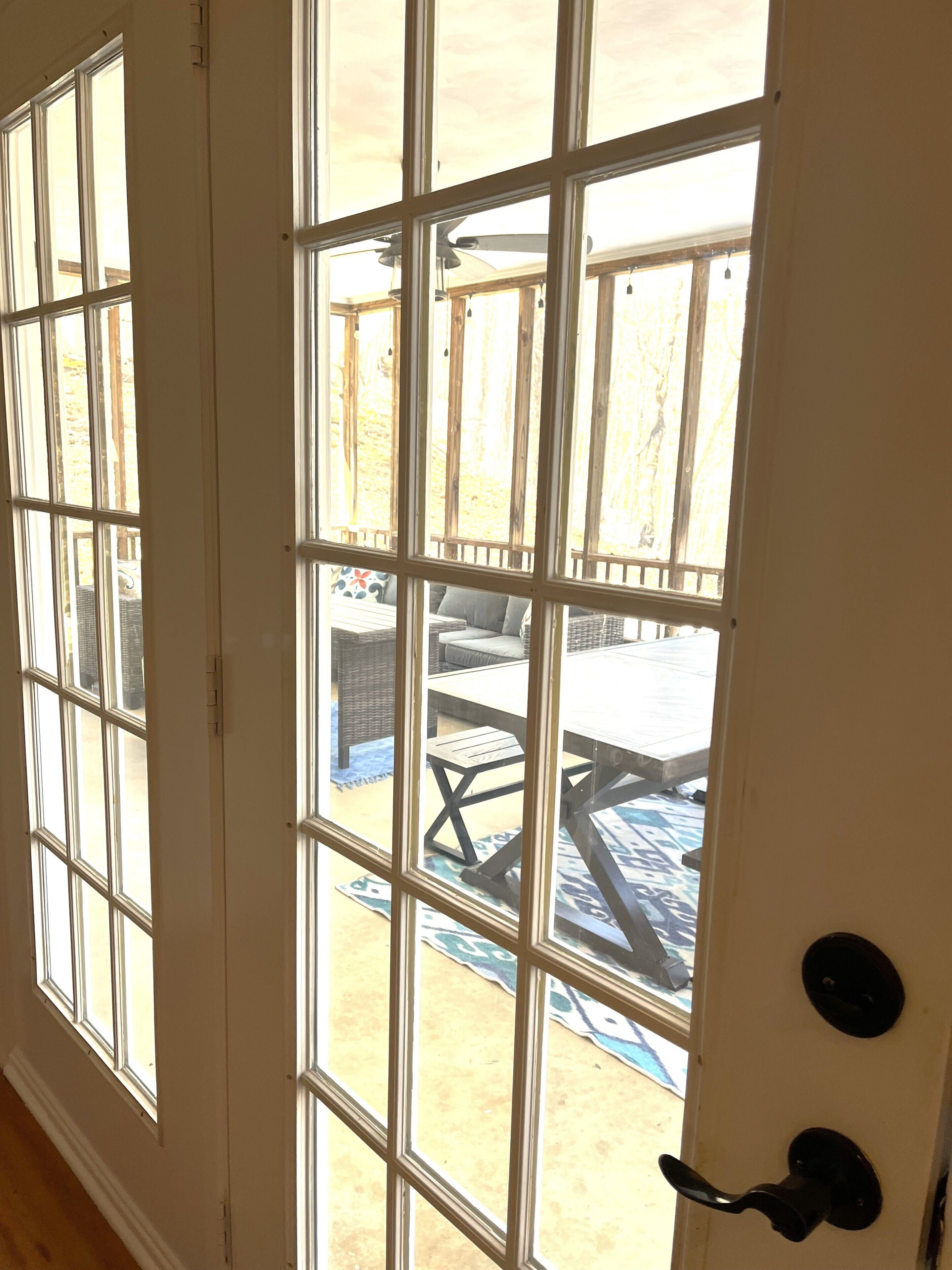 Blick durch ein großes, raumhohes Fenster mit Holzrahmen auf eine Außenpatio mit Tisch und Stühlen.