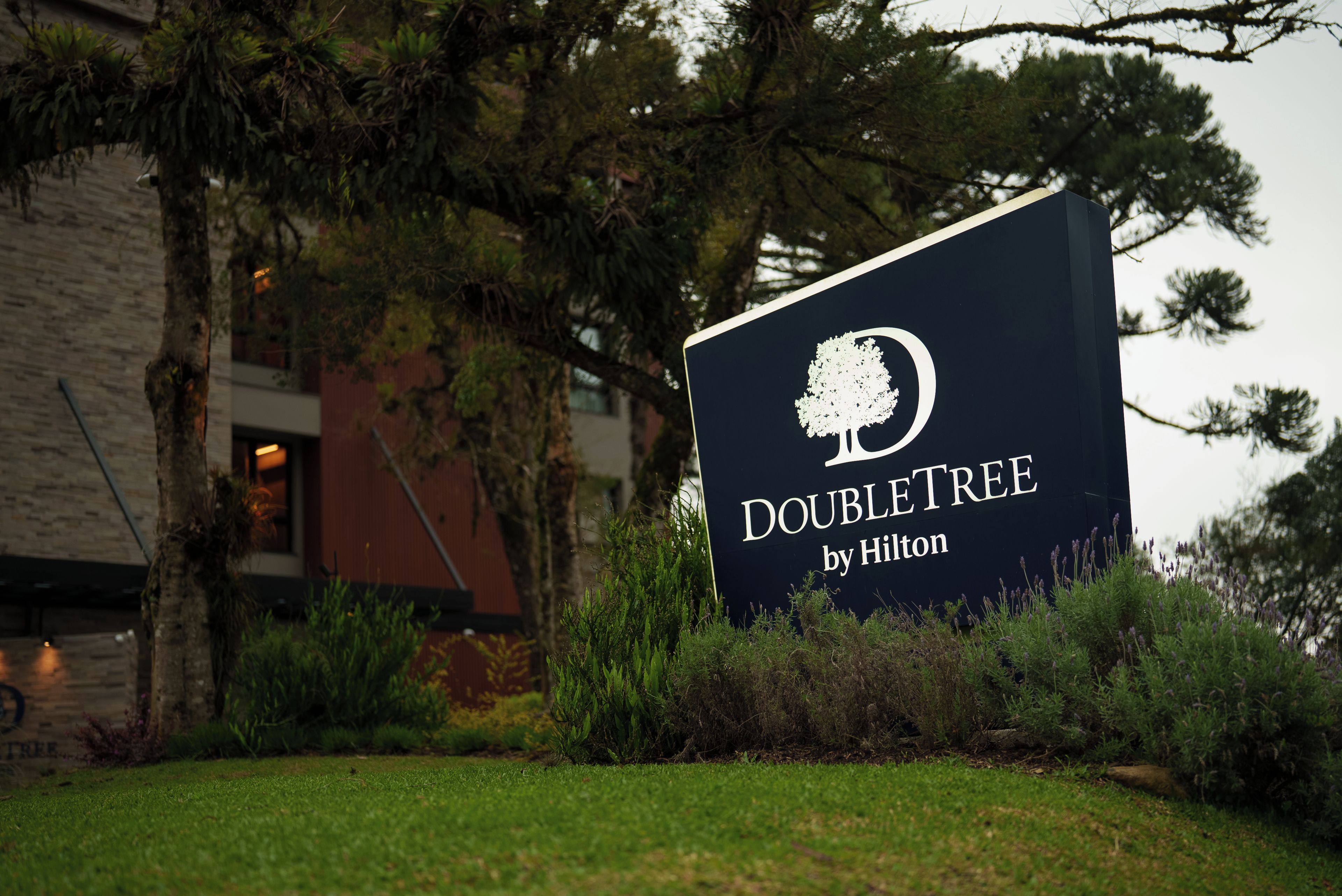 A sign for 'DOUBLETREE by Hilton' with a tree logo, surrounded by greenery and a building in the background.