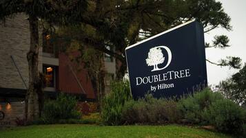 Un letrero de 'DOUBLETREE by Hilton' con el logotipo de un árbol, rodeado de follaje y un edificio al fondo.