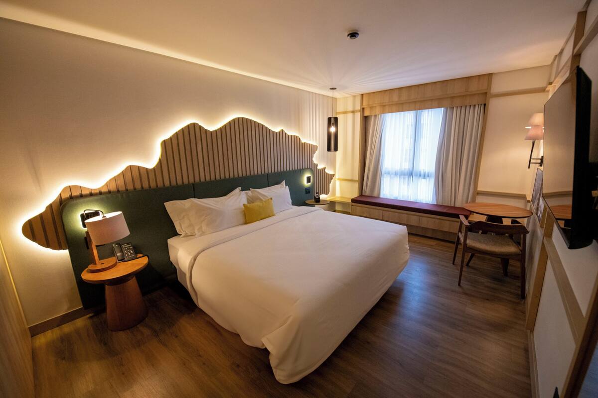A hotel room with a large bed, bedside table with a lamp, a small table with a chair, and a window with curtains.