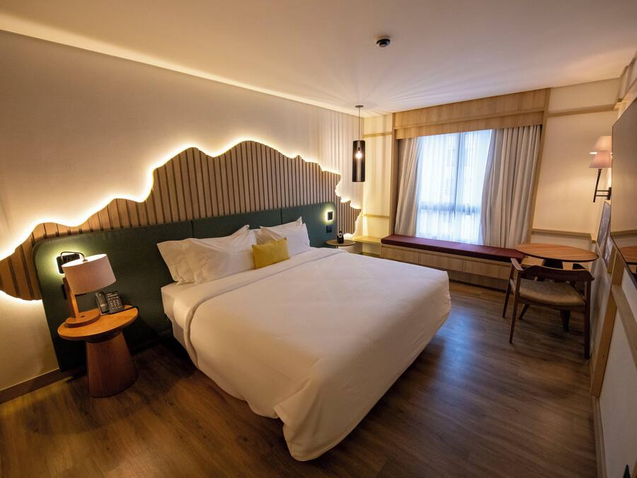 Habitación de hotel con una cama grande, mesita de noche con lámpara, una pequeña mesa con una silla y una ventana con cortinas.