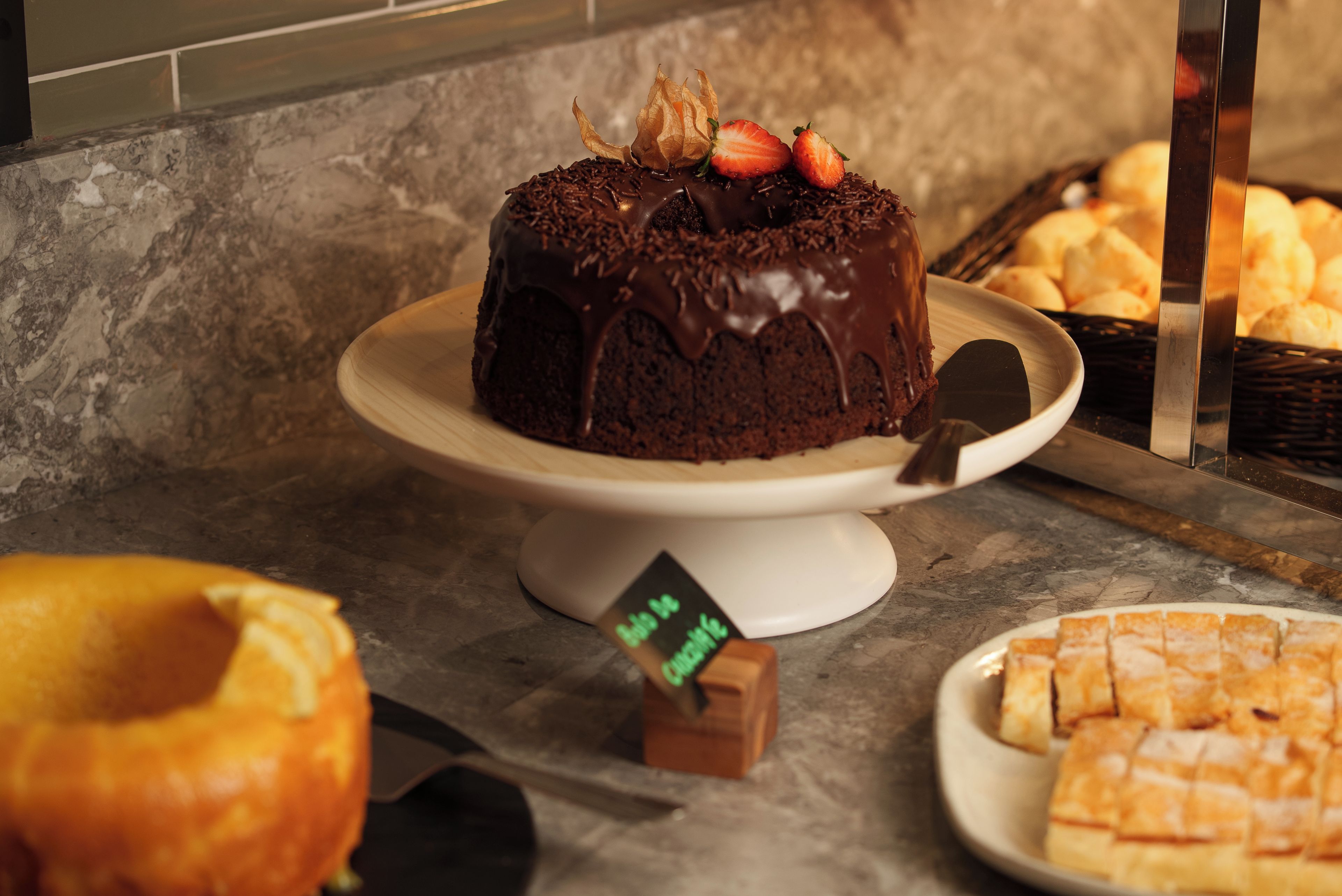 Un pastel de chocolate con fresas sobre un pedestal blanco, rodeado de otras preparaciones horneadas.