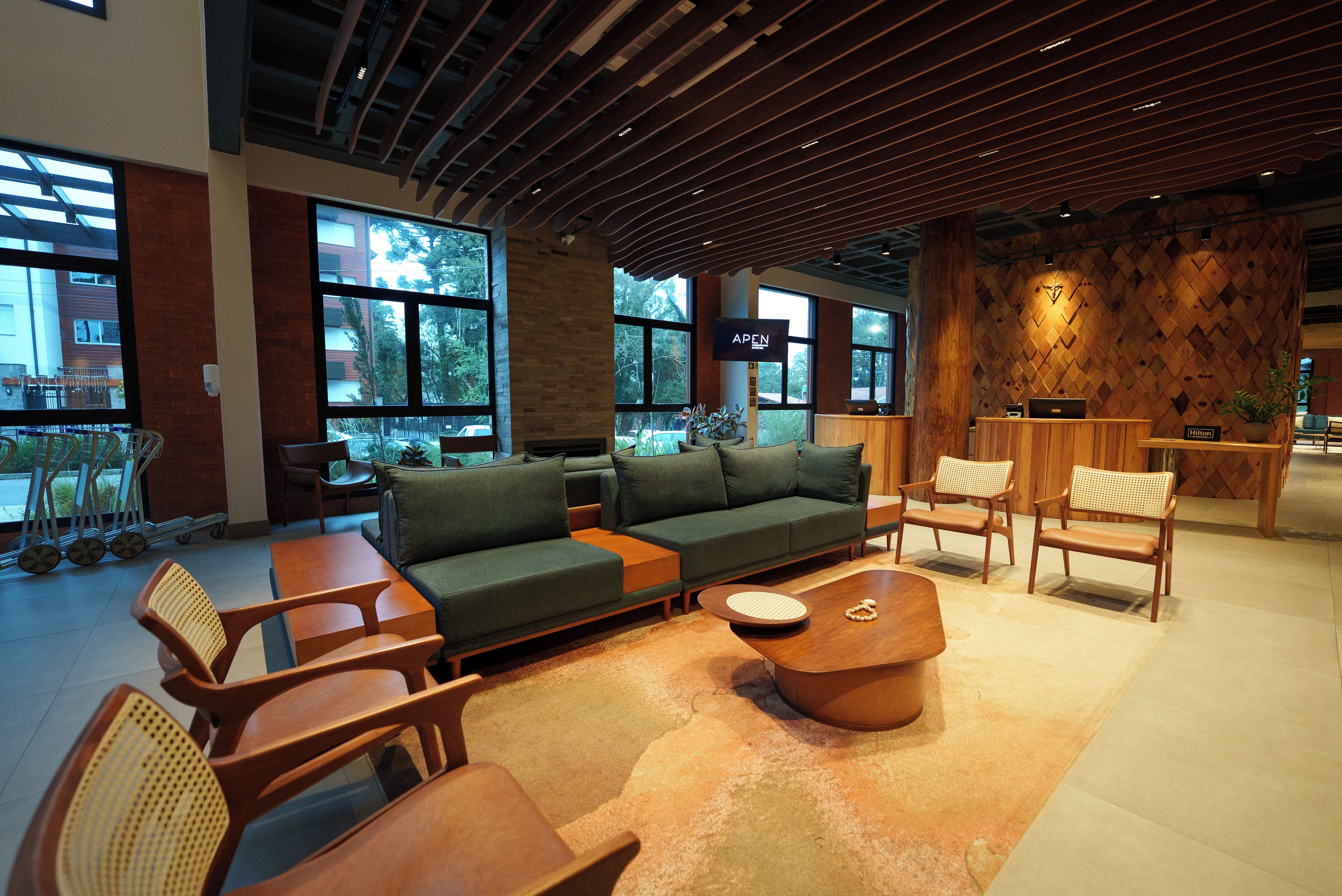 A modern hotel lobby with a green sofa, wooden chairs, a coffee table, and large windows offering views of the outside.