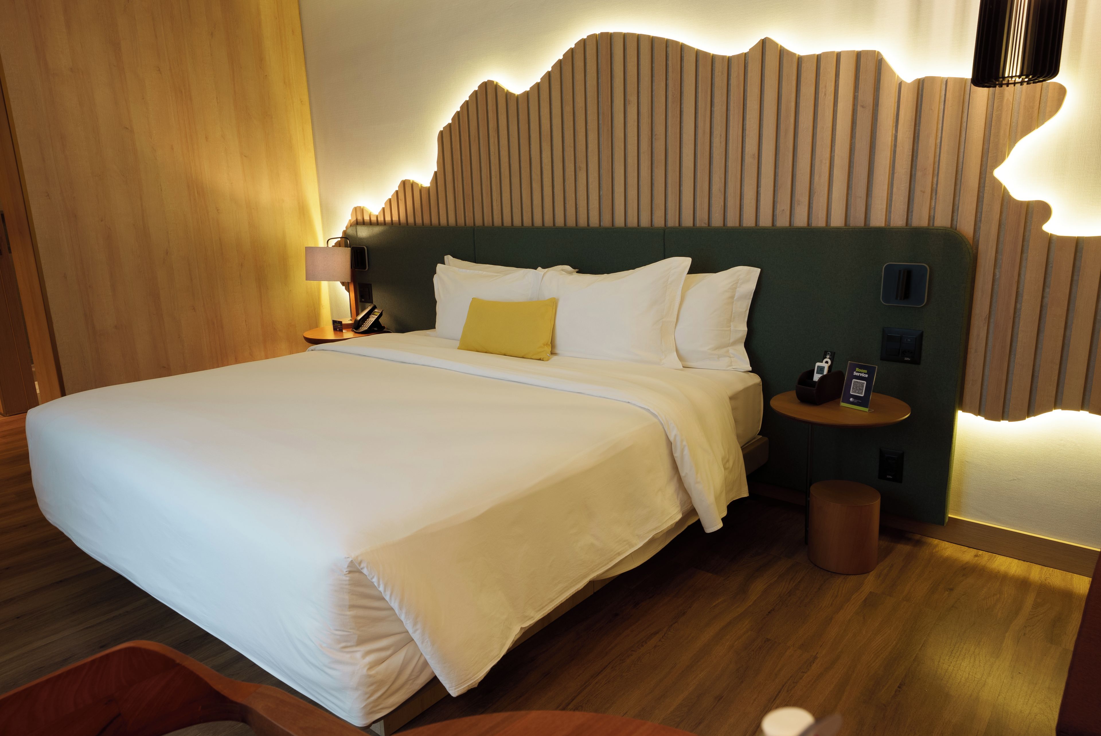 Una habitación de hotel con una cama grande, una mesita de noche con lámpara y un panel de control en la pared.