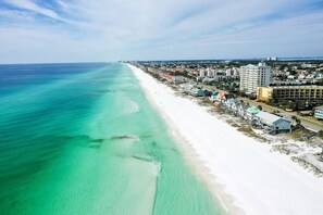 An aerial view of a beachside town with a long stretch of white sand, clear turquoise waters, and a mix of residential and commercial buildings along the shore.