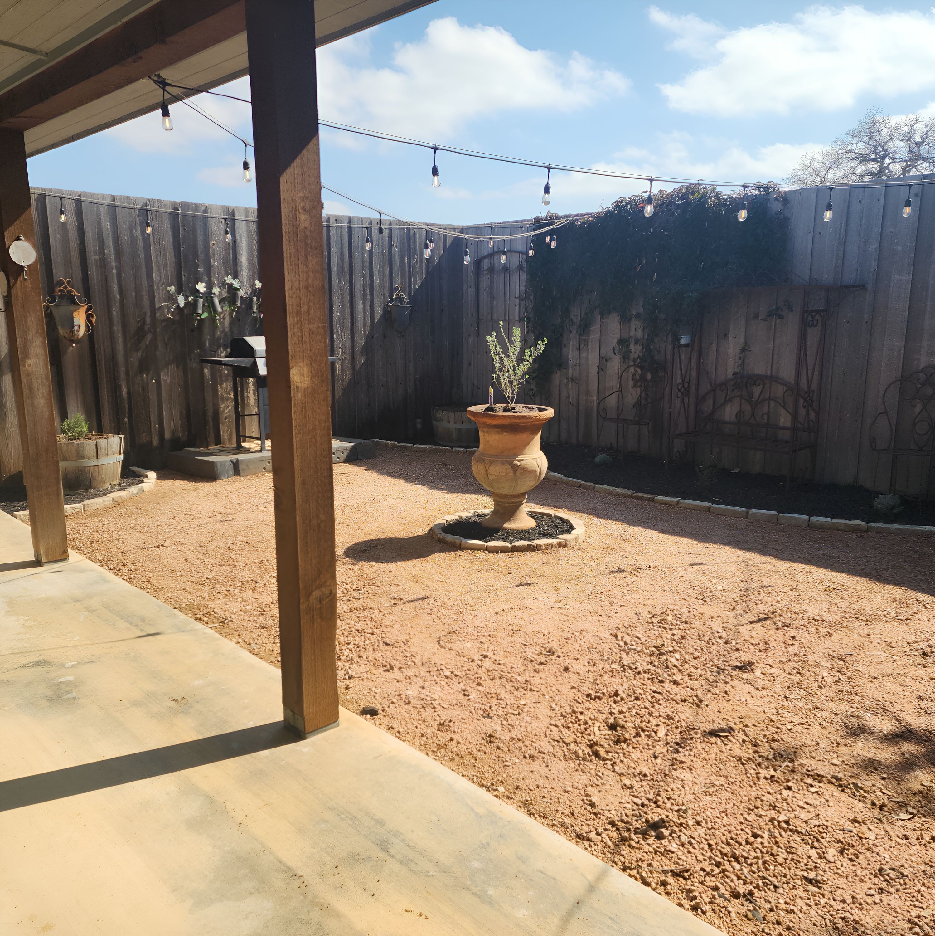 A covered patio area with a gravel ground, a large terracotta pot with a plant, and a wooden fence in the background.