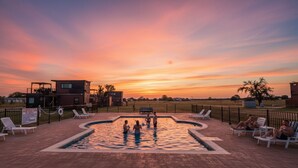 Une piscine avec des chaises longues, une terrasse entourée d’une clôture et des bâtiments en arrière-plan.