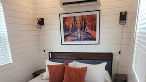 Un lit bien fait, recouvert d’une couette blanche et agrémenté de deux oreillers marron, une photo encadrée représentant un pont accrochée au mur, et une petite table de chevet avec une lampe.