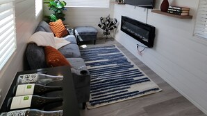 Un salon moderne avec un canapé gris, un tapis à rayures bleues et blanches, un téléviseur à écran plat fixé au mur et une petite table sur laquelle sont posées des bouteilles de vin.