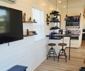 A compact kitchen with white wooden walls, a flat-screen TV, a built-in oven, and a coffee maker. There are two stools and a small shelf with decorative items. - Unit 5 Porto Estacao Stylish Stay (Fredericksburg)