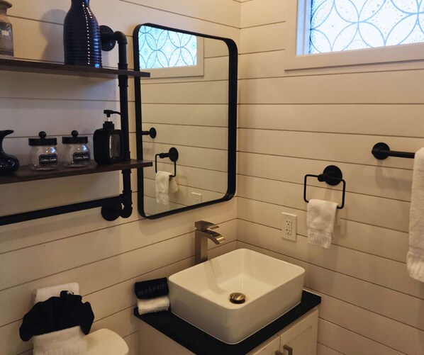 Une salle de bain moderne avec un lavabo blanc, un plan de toilette noir et un grand miroir. On y trouve deux porte-serviettes, une fenêtre ornée d’un motif géométrique et une étagère contenant des articles de toilette.