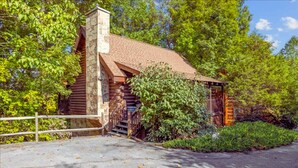 Un chalet rustique en rondins de bois, doté d’une cheminée en pierre, entouré d’une végétation luxuriante et accessible par une allée pavée.