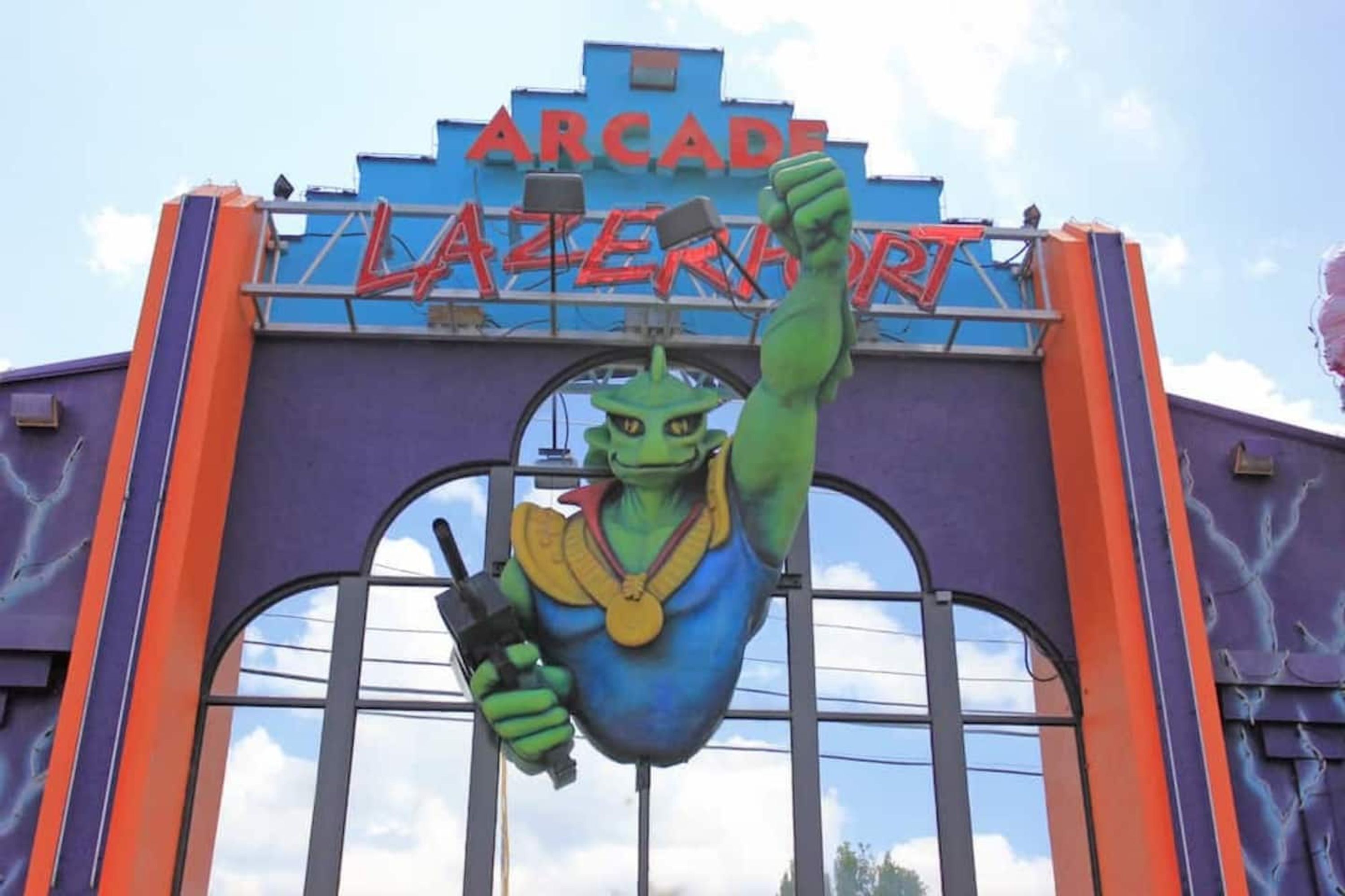 Um mascote alienígena verde com o punho levantado, segurando uma medalha e uma arma, em frente a uma placa que diz 'ARCADE LEMONADE'.