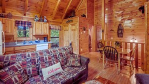 L’intérieur chaleureux d’un chalet avec un plafond en bois, un canapé agrémenté d’un coussin où est inscrit « Bienvenue », une table à manger avec des chaises et un coin cuisine doté de placards et d’une fenêtre.