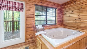 Une cabane avec un intérieur en bois, une grande fenêtre avec des stores, une terrasse en bois à l’extérieur et un jacuzzi.