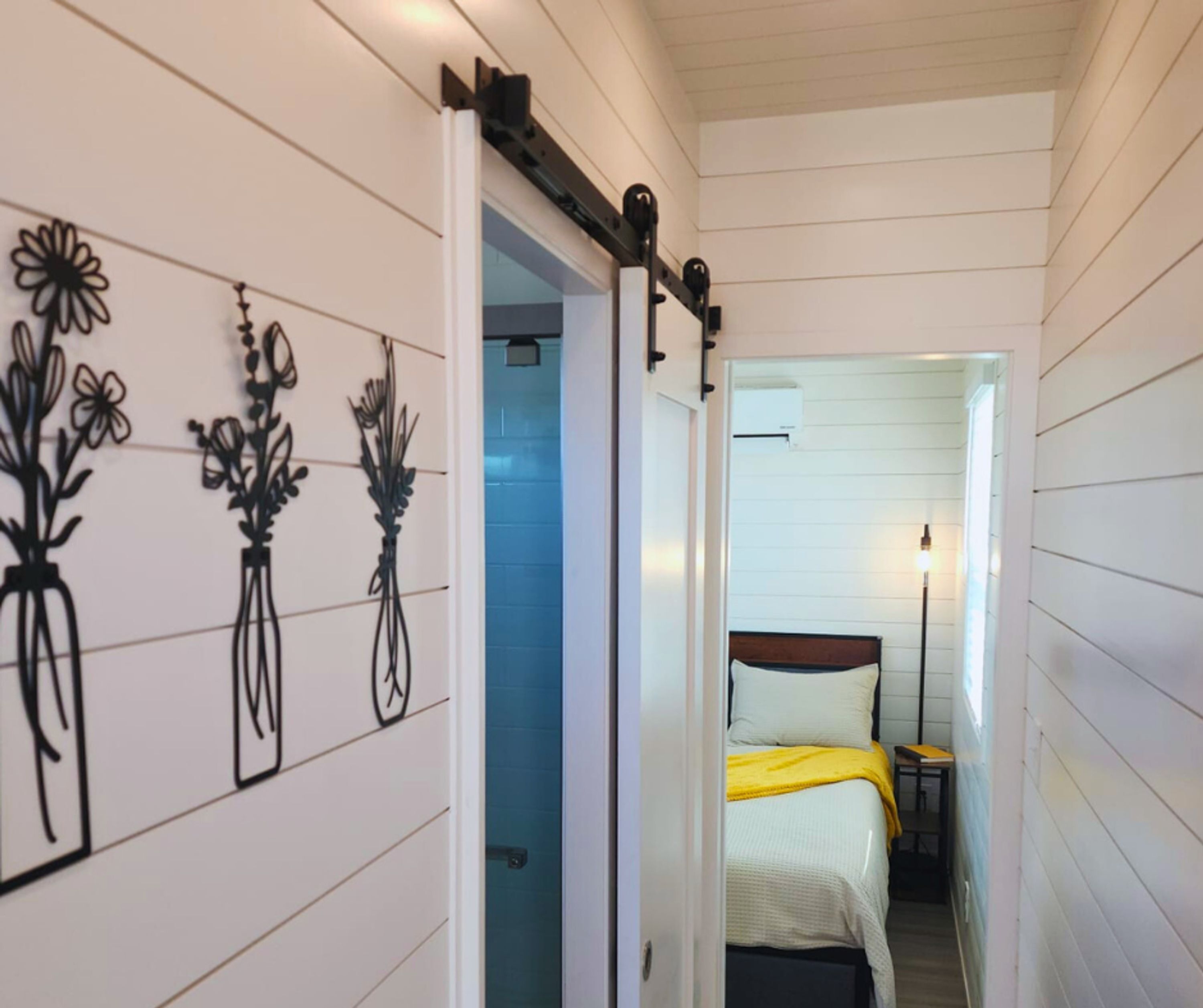 A narrow hallway with a bed, a nightstand, and a lamp. There are wall decorations of flowers.