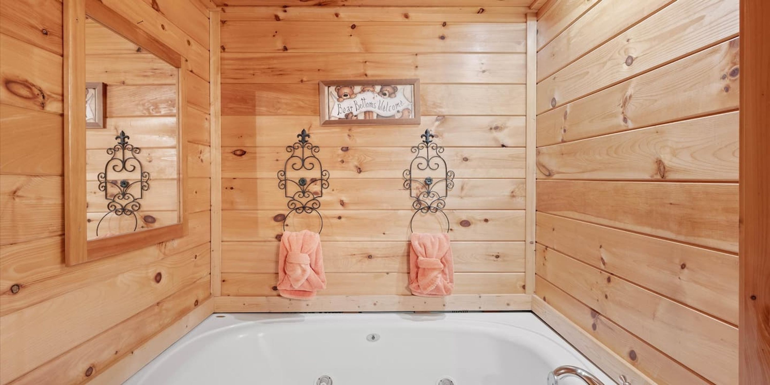A wooden cabin bathroom with a bathtub, two towels hanging on metal towel rings, and a welcome sign above the tub.