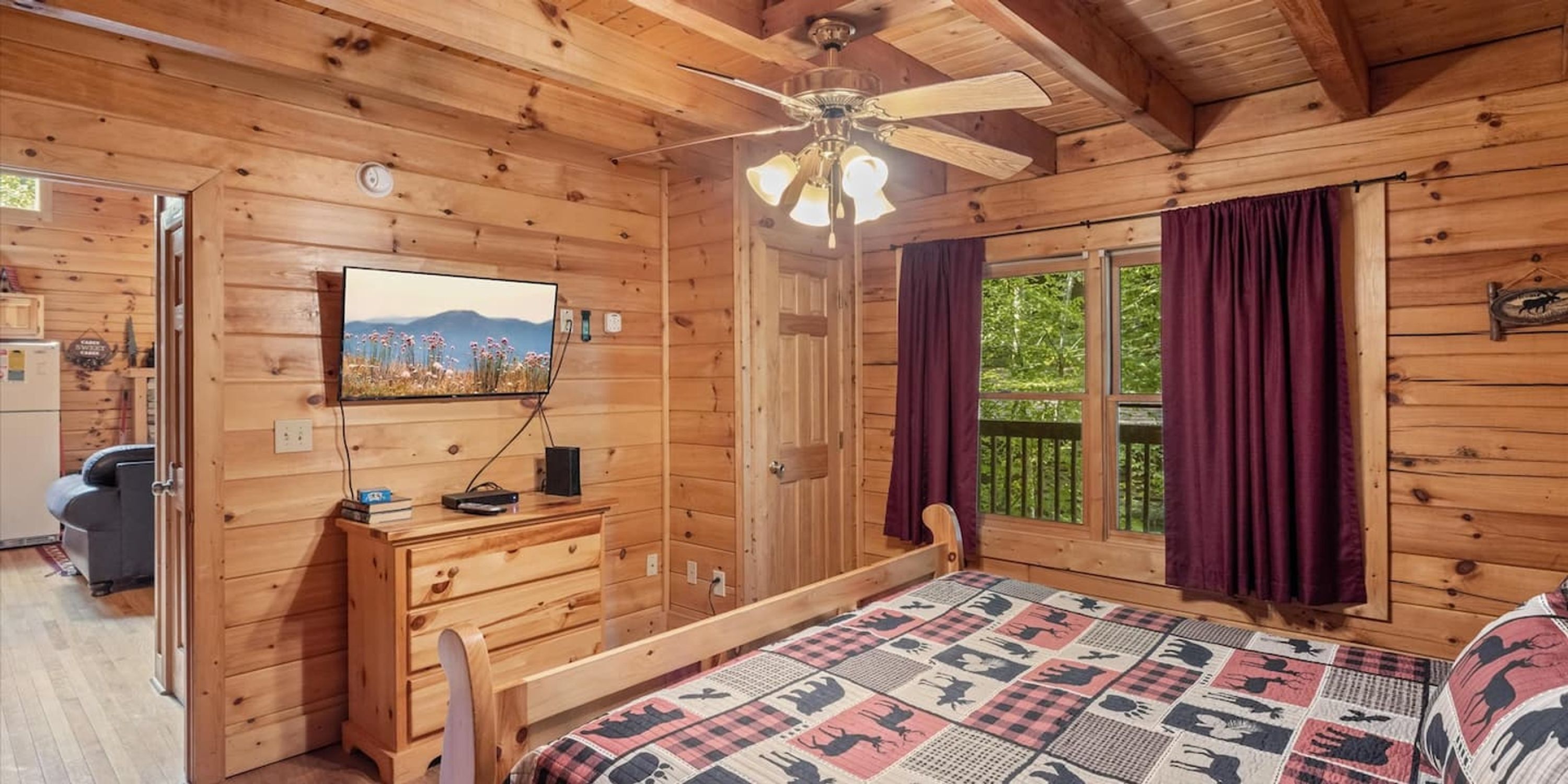 Une chambre dans une cabane en bois, comprenant un lit, un bureau, une chaise, un ventilateur de plafond et une fenêtre avec des rideaux.