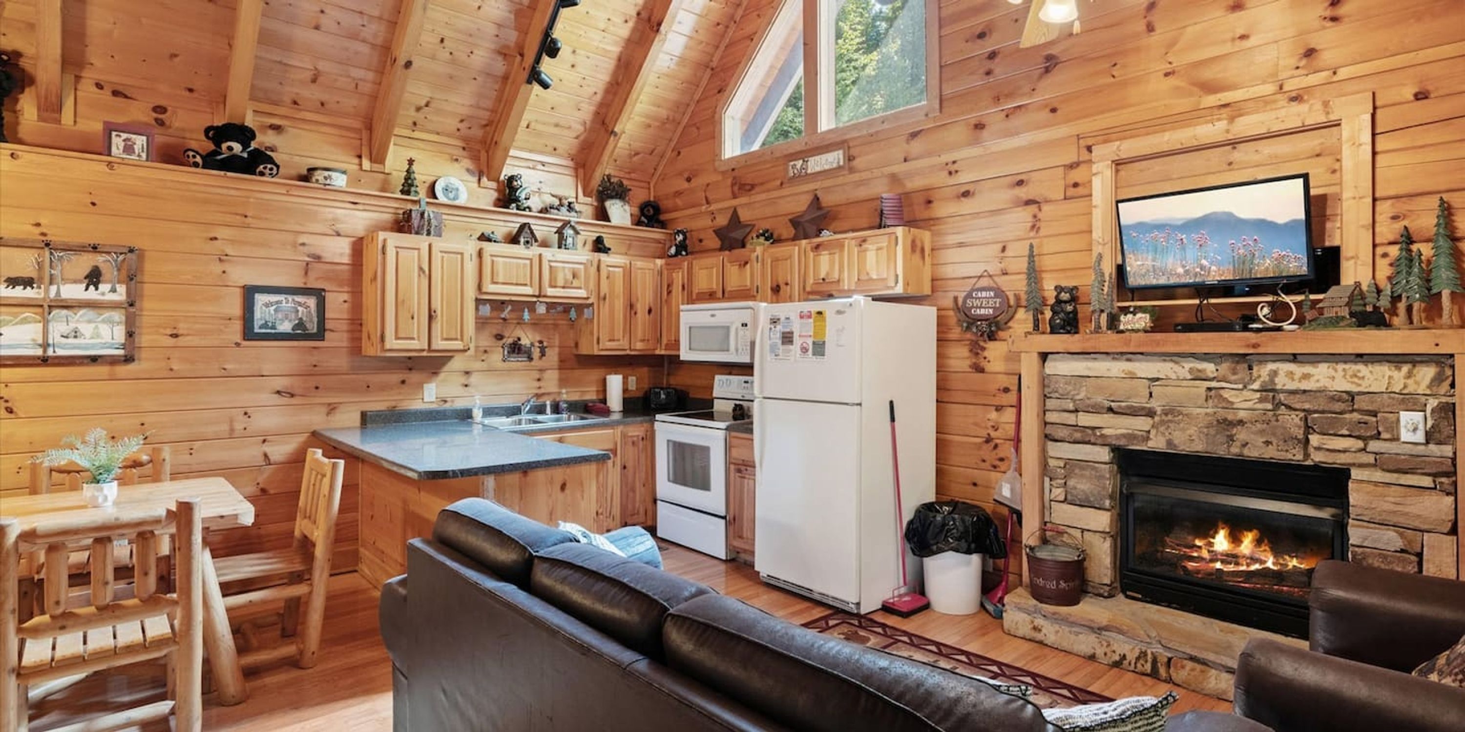 A cozy cabin-style kitchen with wooden walls, a stone fireplace, a dining area with a wooden table and chairs, and a comfortable seating area with a leather couch.