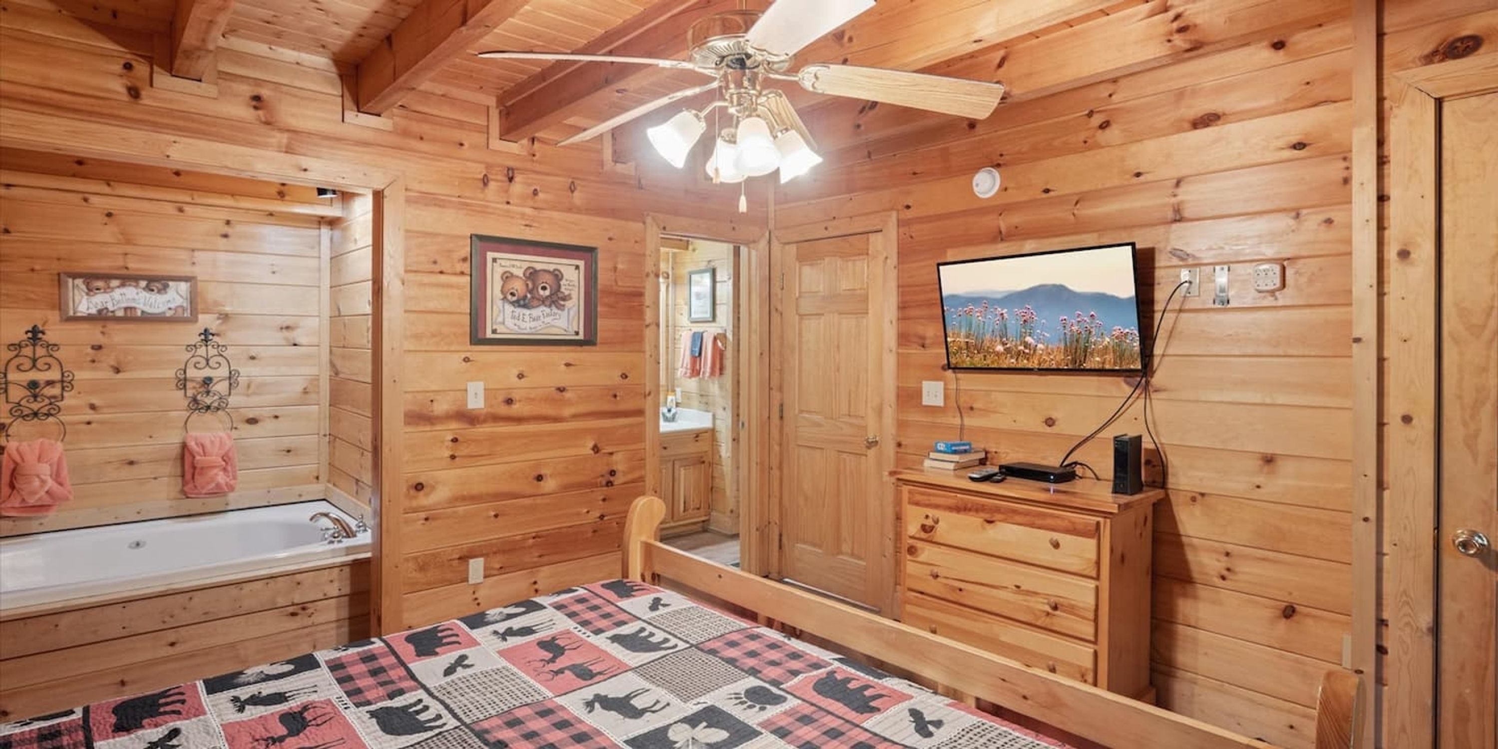 Habitación de cabaña de madera con cama, ventilador de techo, televisor montado, cómoda de madera y baño con lavamanos y toallero.