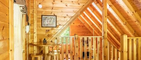 A wooden cabin interior with a high ceiling, a staircase, and a small table with stools.