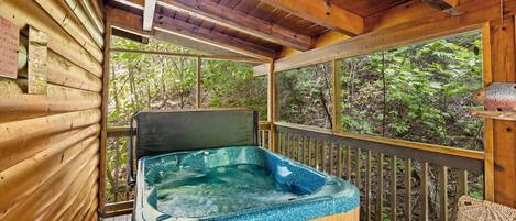 A wooden cabin with a hot tub, large windows, and a view of the forest.