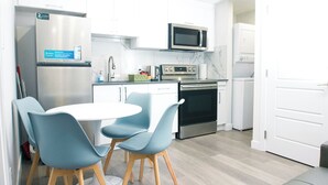 Una cocina moderna con armarios blancos, un refrigerador de acero inoxidable, un microondas, una estufa y una zona de comedor con dos sillas azules y una mesa blanca.