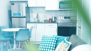 Una cocina moderna con armarios blancos, un refrigerador de acero inoxidable, un microondas y un horno. Hay una mesa de comedor con sillas azules y un cojín celeste con lunares en el sofá.