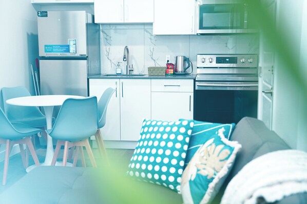A modern kitchen with white cabinets, a stainless steel refrigerator, a microwave, and an oven. There is a dining table with blue chairs and a teal polka dot pillow on the sofa.