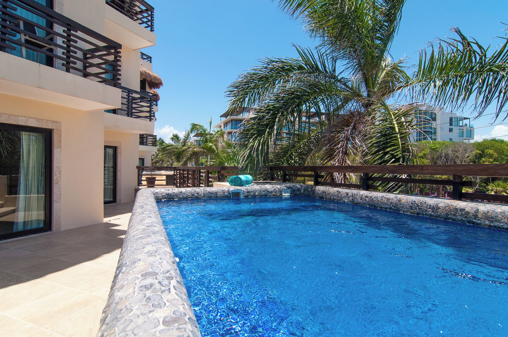 A swimming pool with stone tiles, surrounded by a wooden deck and palm trees.