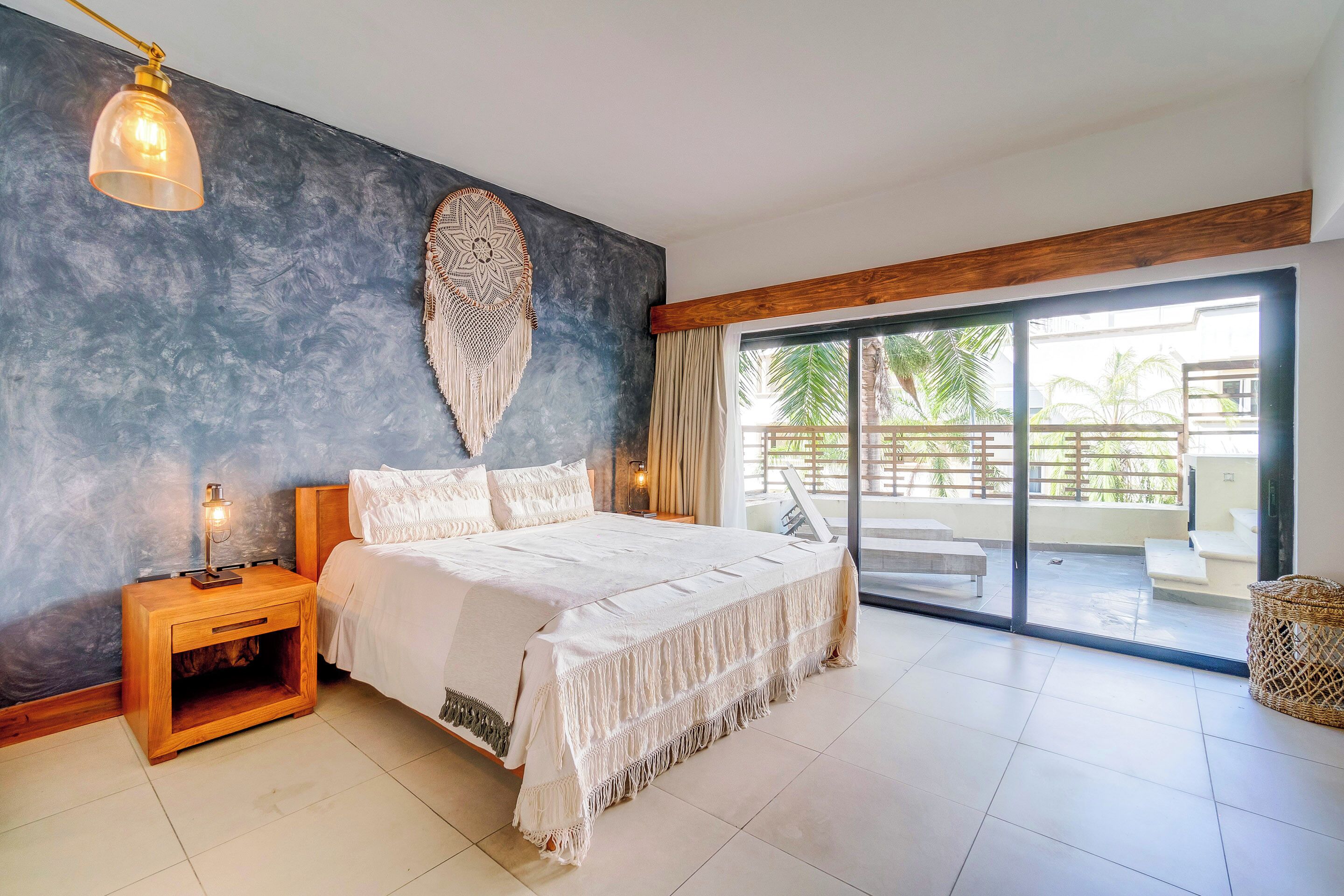 A bedroom with a large bed, a nightstand, a wall hanging, and a view of the outside balcony.