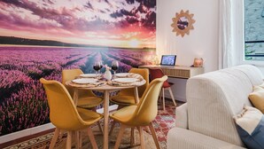 Een eethoek met een tafel voor vier personen, een laptop op een bijzettafeltje, en een muurschildering van een zonsondergang.