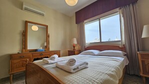 A hotel room with a bed, a wooden dresser, a mirror, a window with curtains, and an air conditioning unit. - 74 LCA - Stay Sea Gate Apartment (Larnaca)