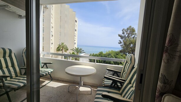 A balcony with striped chairs and a small table, overlooking a building and a body of water. - 74 LCA - Stay Sea Gate Apartment (Larnaca)